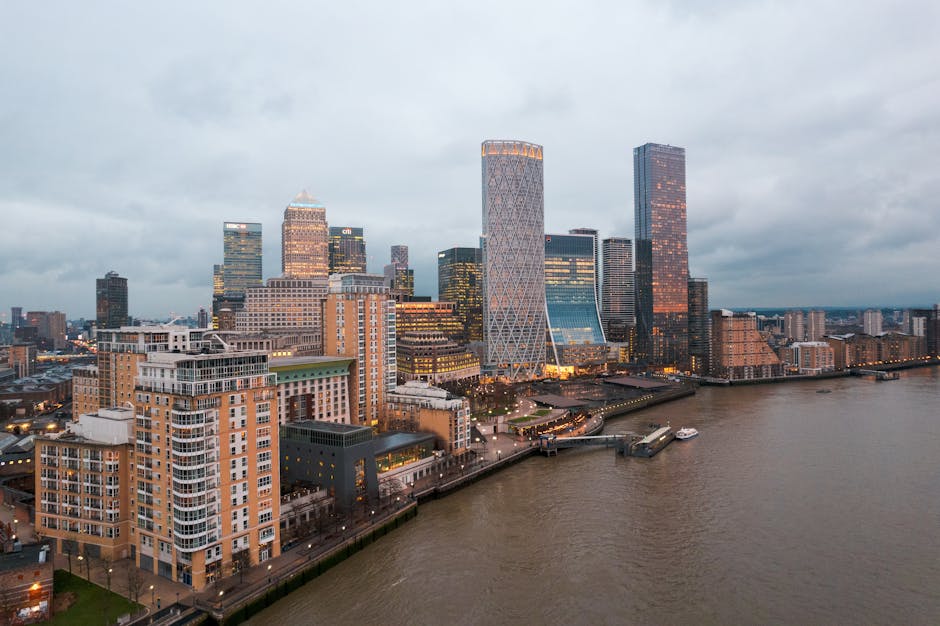 A panoramic view of London's Canary Wharf district from across the River Thames, featuring modern glass skyscrapers, including buildings branded with HSBC and Citibank, under a clear blue sky. The sce