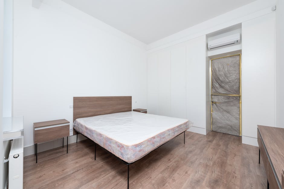 A neatly made bedroom with a beige upholstered bed against a cream-colored wall. Above the bed, there is a decorative round mirror surrounded by an abstract sunburst mirror design with various sized c